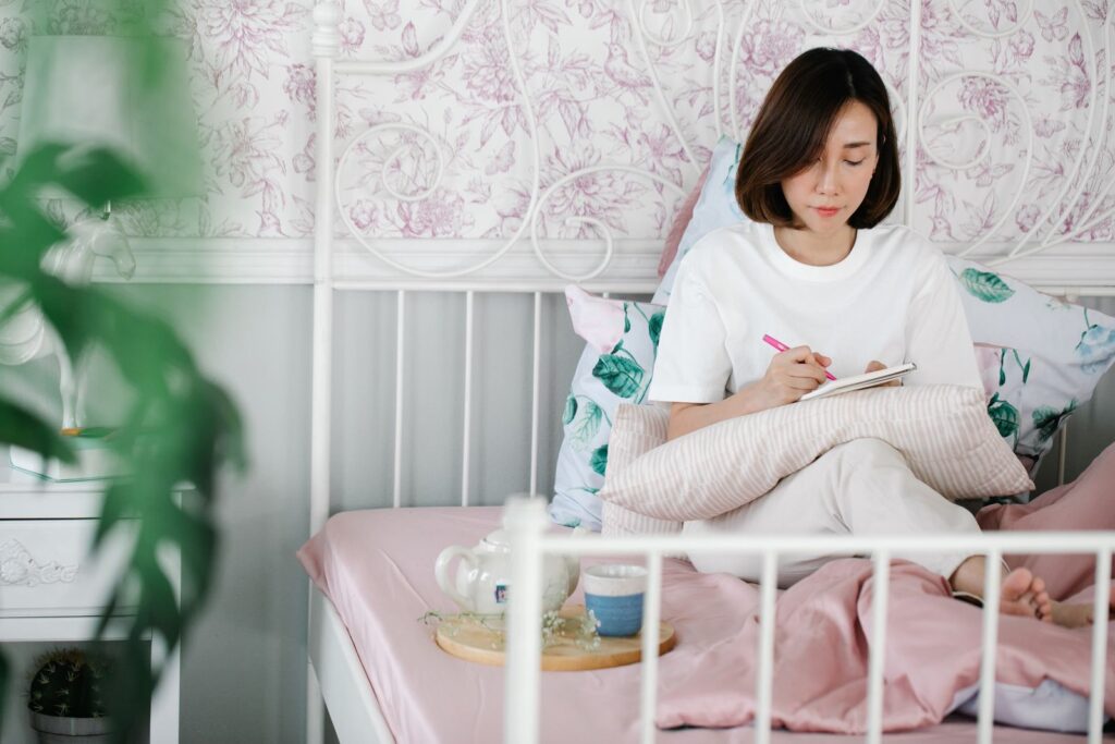 Mulher sentada na cama escrevendo em um diário, em um quarto sereno e aconchegante — foto: Pexels