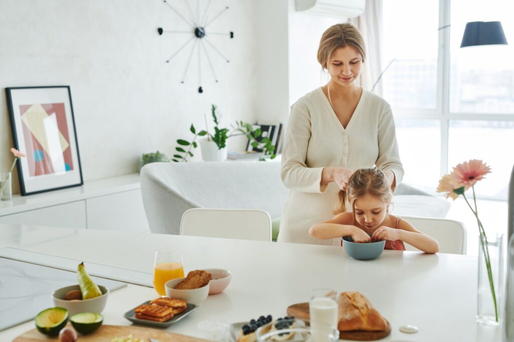 Mãe trançando o cabelo da filha em uma cozinha clara, cercada por itens de café da manhã — foto: Pexels