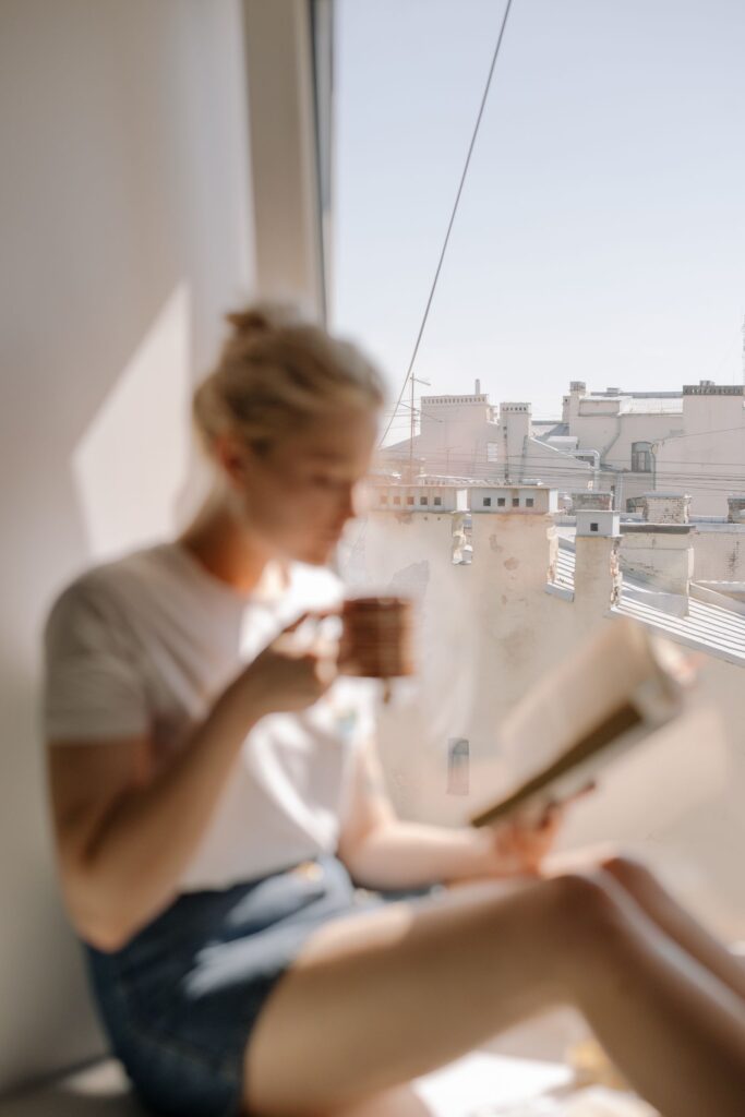 Mulher sentada perto da janela, lendo e tomando um café, com vista para telhados em um dia claro — foto: Pexels