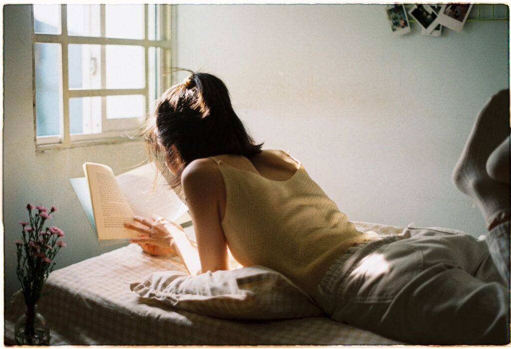 Mulher deitada lendo um livro perto da janela com luz da manhã — foto: Pexels