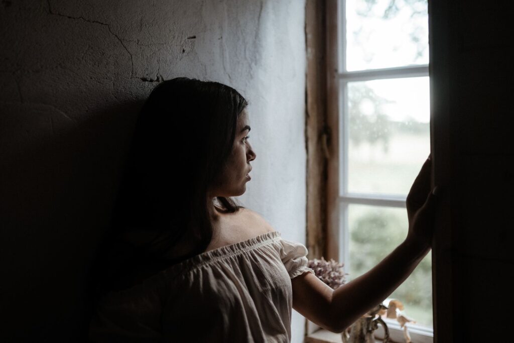 Mulher olhando pensativa pela janela, luz suave — foto: Pexels