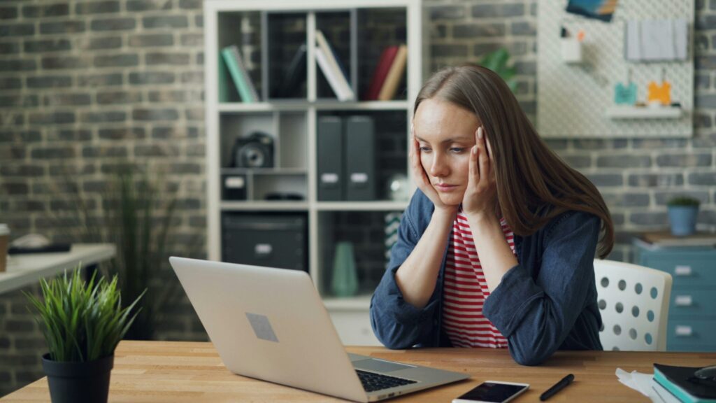 Mulher apoiando a cabeça nas mãos, olhando cansada para o laptop na mesa de trabalho