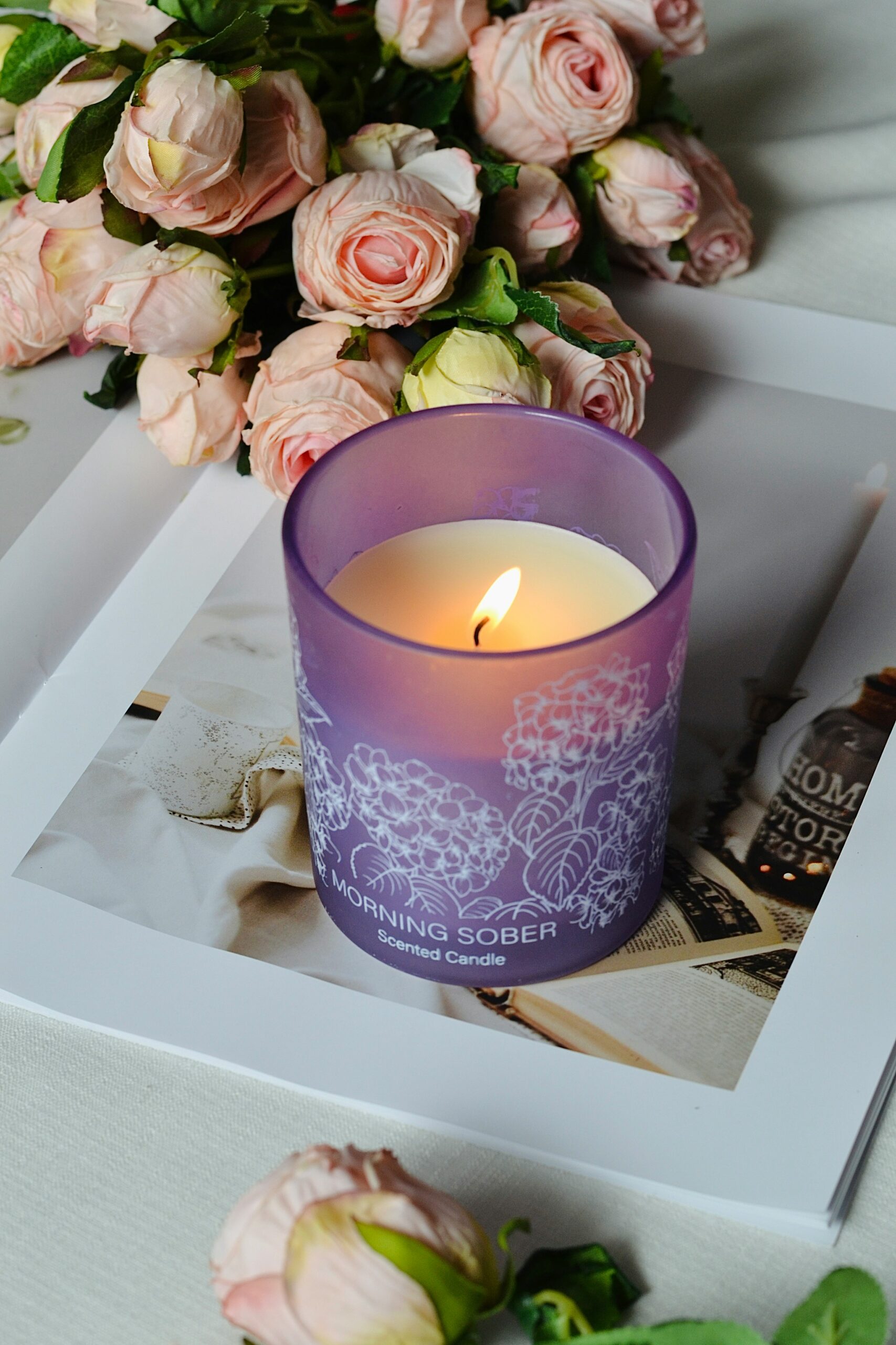 Vela aromática acesa em copo roxo ao lado de flores rosa