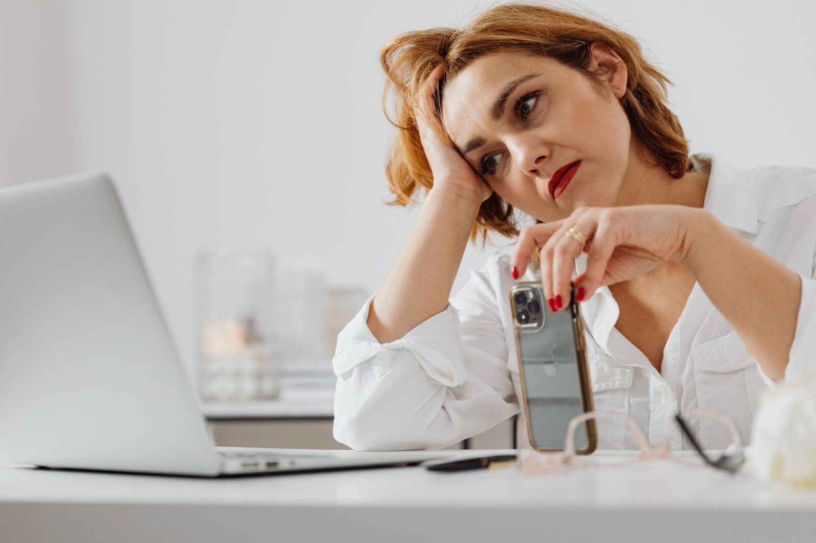 Procrastinação: mulher pensativa segurando o celular diante da mesa de trabalho