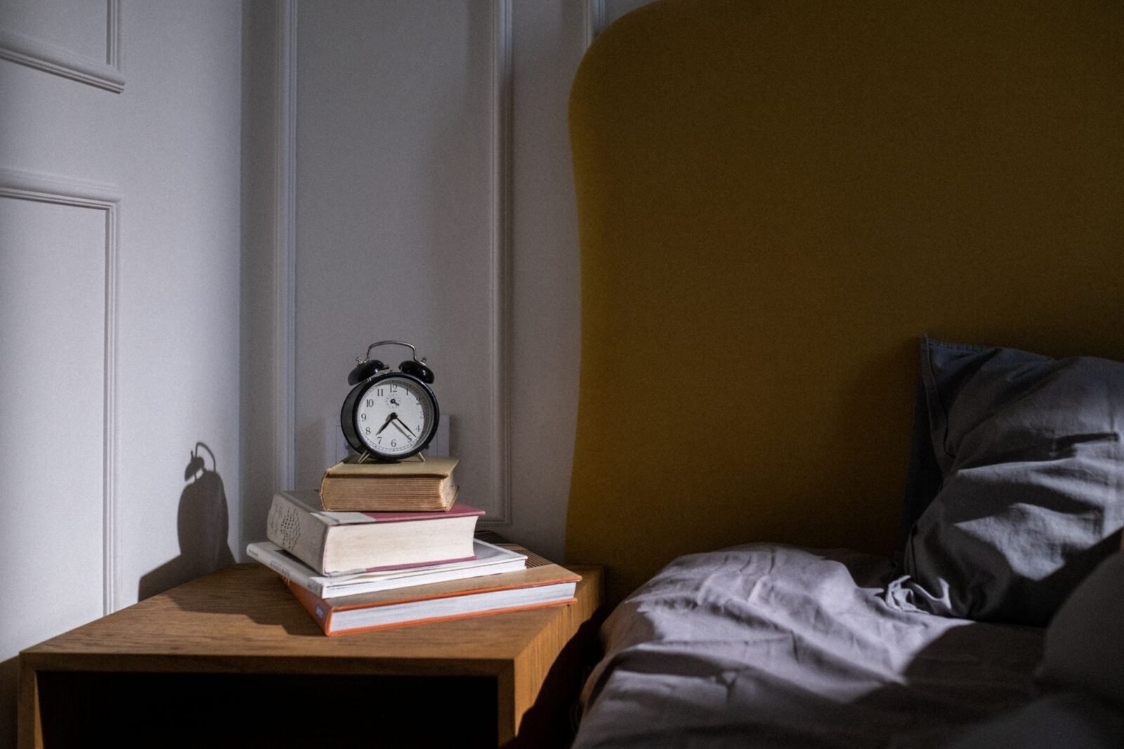 Despertador analógico sobre uma pilha de livros na mesa de cabeceira, ao lado da cama, sem celular por perto