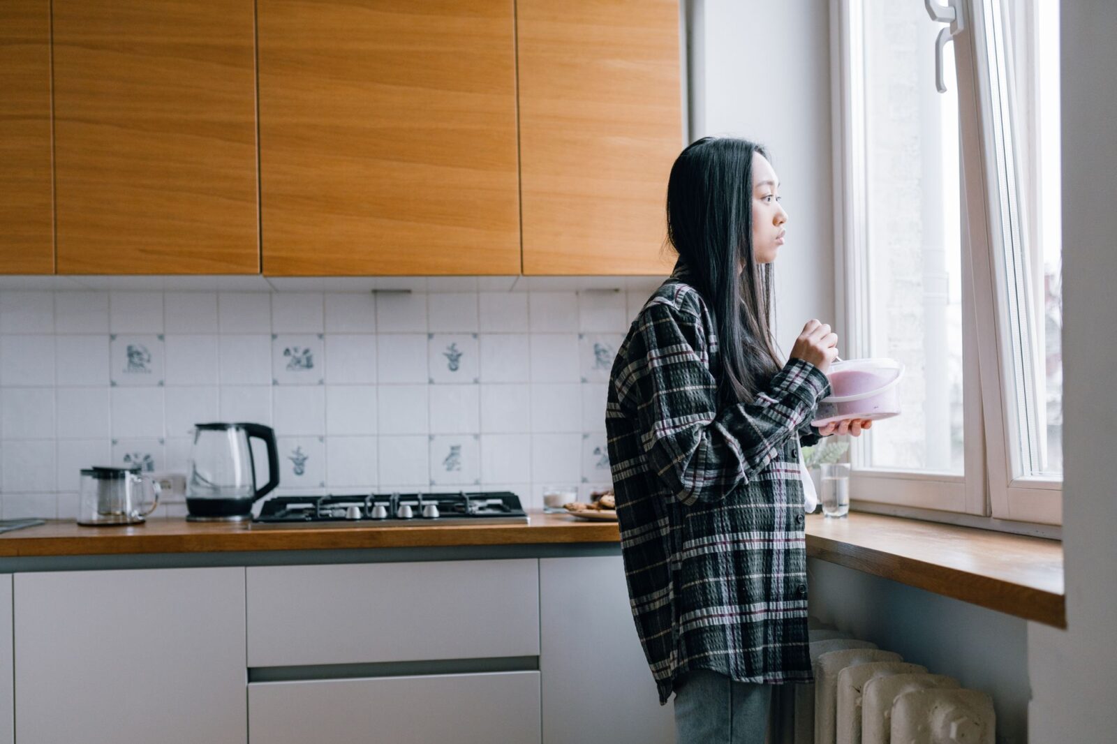 Mulher na cozinha segurando um doce e olhando pela janela, imagem sobre fome emocional