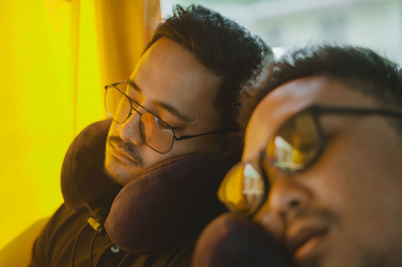 Dois homens de óculos dormindo sentados num ônibus, com almofadas de pescoço em formato de U